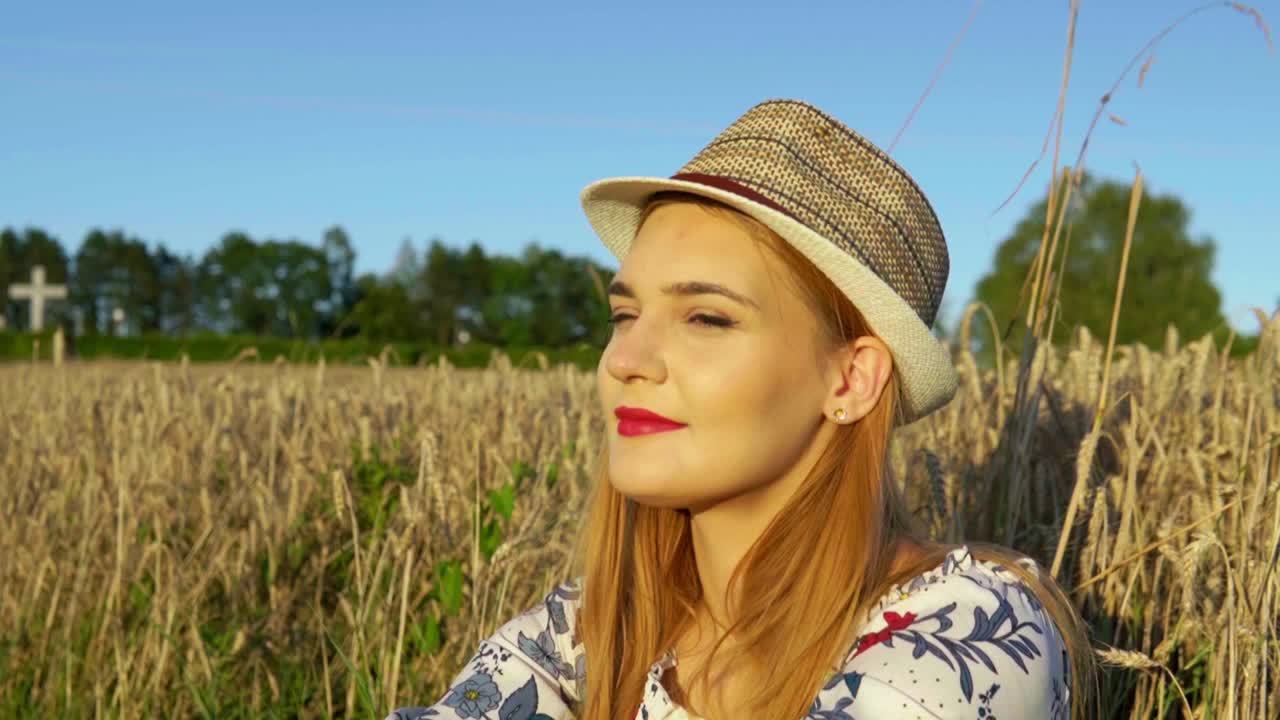 Stunning HD footage of a white caucasian girl in a dress with a knitted hat and red lipstick sitting in a wheat field, lost in thought, basking in the setting sun's warm embrace