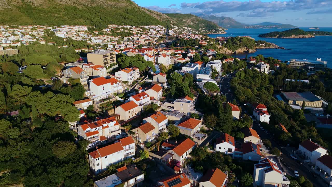 Aerial close-up drone circles Dubrovnik hillside homes, highlighting terracotta red roofs and narrow streets, with the golden sunset casting warm light over the historic town and surrounding landscape