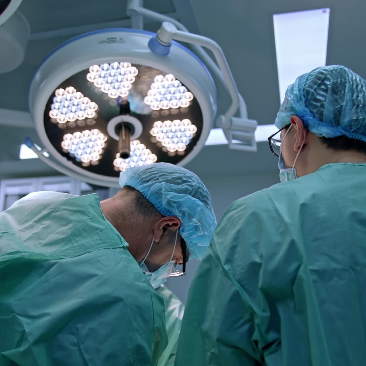 Rear view of two male doctors working at operational table. Uniformed professional perform procedure. Round lamps are at backdrop