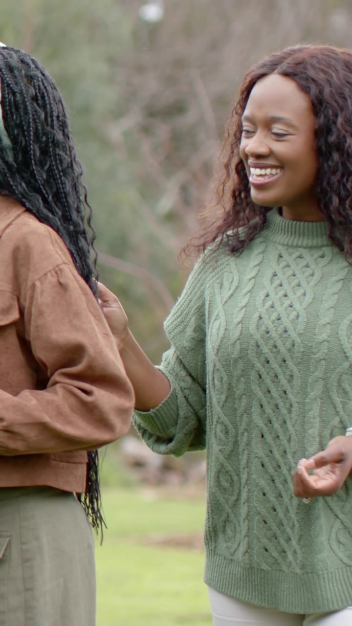 Vertical video: Smiling woman in green knit reaching and greeting friend by touching braid at park