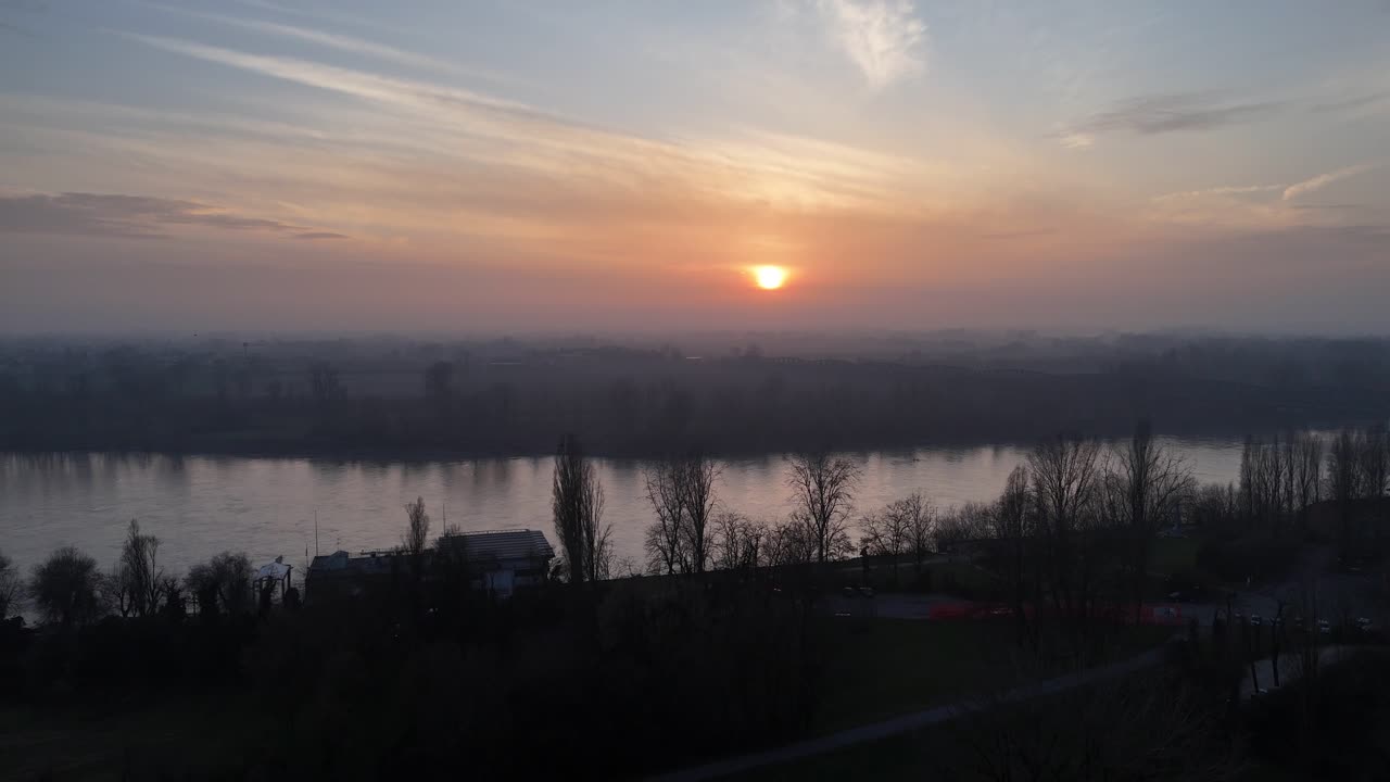 Flygvy av solnedgång över Po-floden vid Cremona i gyllene timmen-belysning, silvrigt grå vattenyta, kala lövträd under vintern och atmosfärisk vinterdimma över Padanas slättlandskap, Lombardiet, Italien