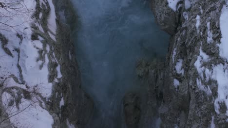 Emerald-water-of-mountain-river-streaming-between-frozen-rocks-covered-by-snow