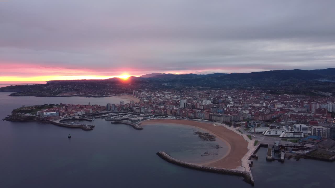 Vista panoramica aerea del paesaggio urbano di Gijón durante un tramonto epico, con spiaggia, edifici del centro città e porto con le montagne sullo sfondo.