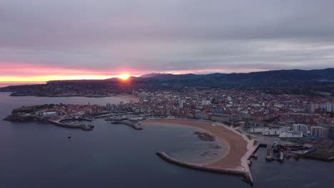 Vista-Panorámica-Aérea-Del-Paisaje-Urbano-De-Gijón-Durante-La-épica-Puesta-De-Sol,-El-Edificio-Del-Centro-Urbano-De-La-Playa-Y-El-Puerto-Con-Montañas-En-El-Fondo