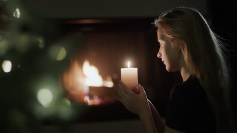 una adolescente sostiene una vela encendida en sus manos. se sienta junto al árbol de navidad, al fondo arde un fuego en la chimenea. vista lateral