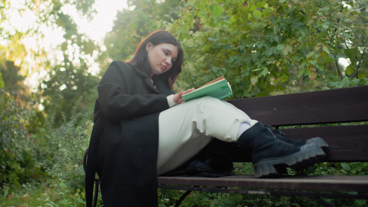 Lower view of free spirited girl seated on park bench with legs up reading book, adjusting hair between pages, paper cup on ground, black coat and boots, gentle sunlight through green foliage