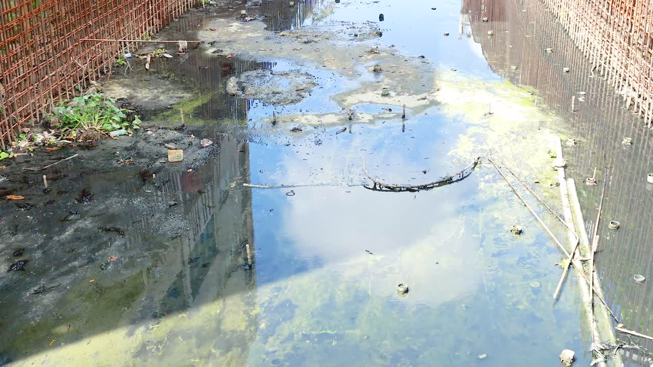 A polluted canal in Phuket's Old Town with stagnant water, debris, and reflections under bright daylight