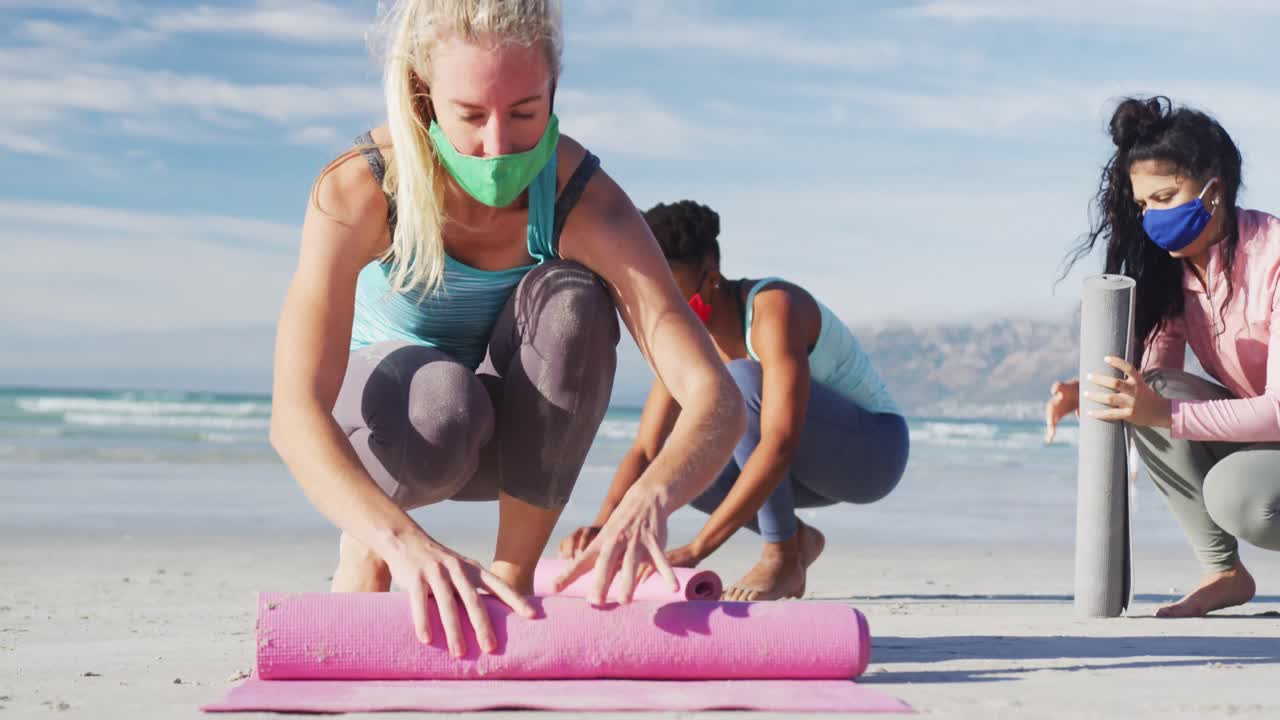 grupo de diversas amigas con máscaras faciales enrollando esteras de yoga en la playa