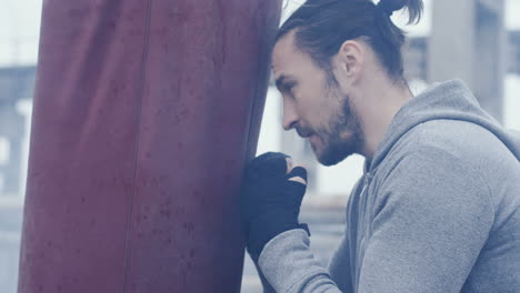 Vista-De-Cerca-De-Un-Hombre-Caucásico-Con-El-Pelo-Largo-Golpeando-Un-Saco-De-Boxeo-Al-Aire-Libre-En-Una-Fábrica-Abandonada-En-Una-Mañana-Nublada