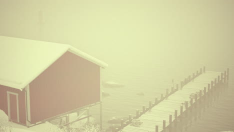 a red wooden house and a wooden dock are in the water on a foggy winter day