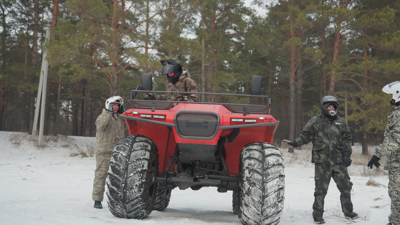 Pilota con casco alla guida di un ATV rosso con l'equipaggio che assiste sulla neve, rapido passaggio di consegne tra i piloti, test dell'acceleratore, rombo del motore, controllo della trazione su sentiero ghiacciato, pini, abbigliamento mimetico tattico, di supporto