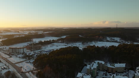 Aerial-View-Over-Woodland-Trees-And-Snow-Covered-Town-Of-Dabrowa-In-Gdynia-During-Golden-Hour-Sunset-On-Horizon