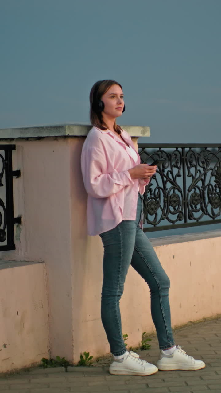 Woman standing outdoors at dusk, leaning against a decorative iron fence, wearing headphones and holding a phone, with soft evening lighting and moon visible in the background