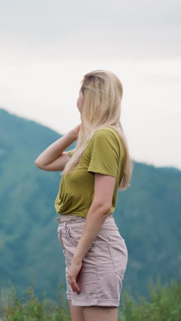 smiling pretty blonde woman amazed of picturesque mountains view touches long loose blonde hair at highland resort on gloomy day slow motion