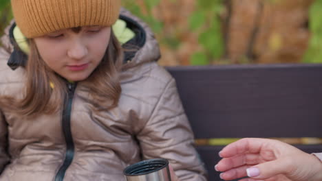 close up of mother hand in beige jacket offering metal cup with warm drink to daughter seated beside her on wooden bench during chilly autumn day, surrounded by golden foliage and soft light