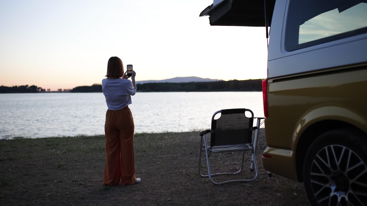 Young Woman Taking Photo of Sunset by Lake with Camper Van