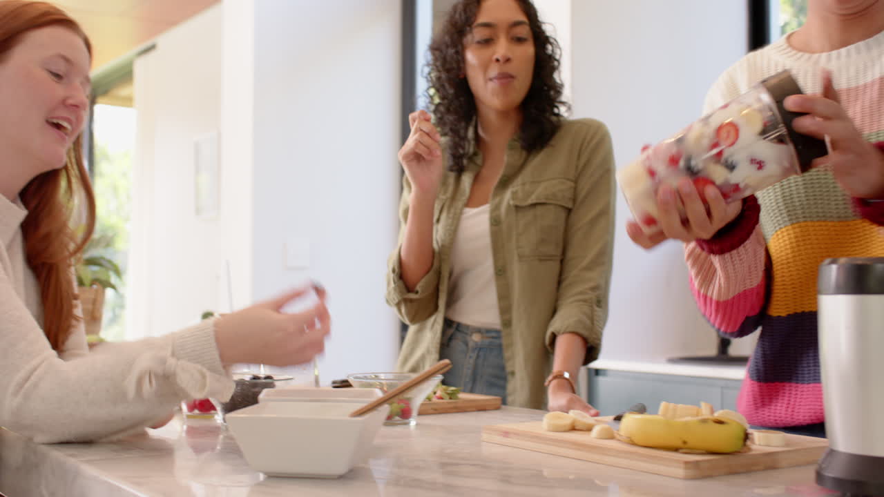 Preparing smoothie, diverse female friends enjoying healthy snacks in kitchen together