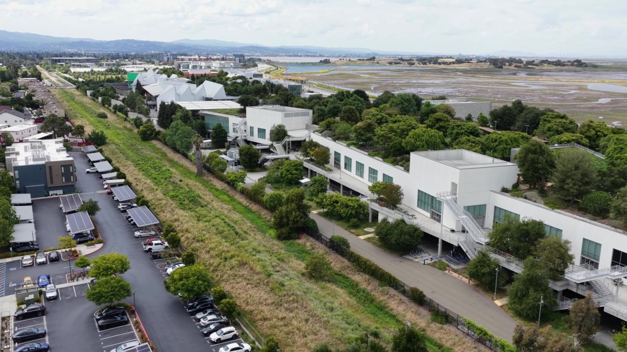 Campus da sede da Meta em Menlo Park com edifícios de escritórios brancos modernos, estacionamento coberto por painéis solares, árvores verdes exuberantes, pântanos da baía e cordilheira sob céu nublado. Califórnia, EUA. Órbita aérea.