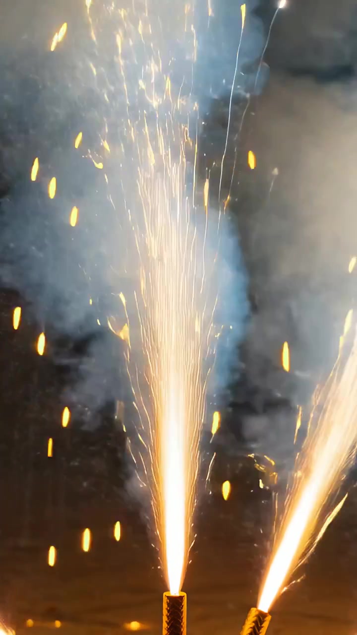 Video verticale: Due fontane pirotecniche accese a terra che emettono scintille e fumo di notte.