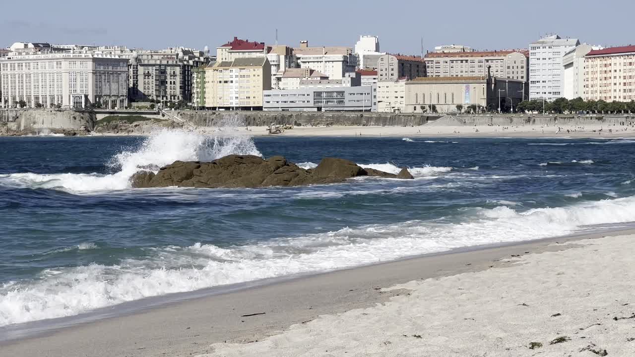 Una foto panoramica della spiaggia di La Coruña in Spagna, che mostra le onde che si infrangono sulla costa rocciosa con alti edifici sullo sfondo.