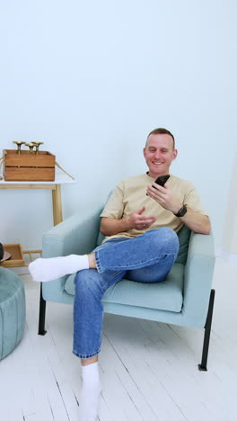 positive smiling caucasian mid-aged man sits in the armchair. man sitting indoors with phone in hands. vertical video.