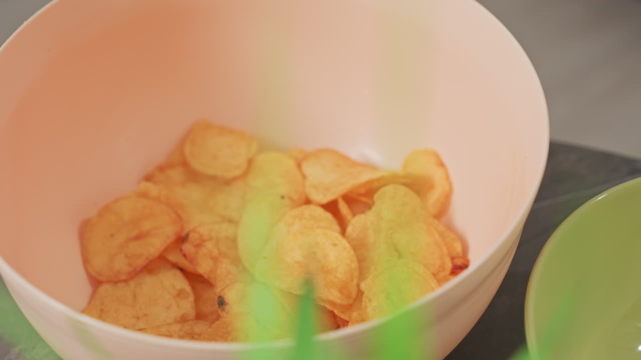 close up of hand taking fried chip from peach colored bowl with golden crispy chips inside, soft focus green plant nearby, casual snack moment, indoors, natural lighting, relaxed environment