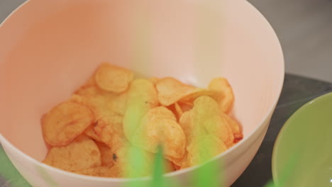 close up of hand taking fried chip from peach colored bowl with golden crispy chips inside, soft focus green plant nearby, casual snack moment, indoors, natural lighting, relaxed environment
