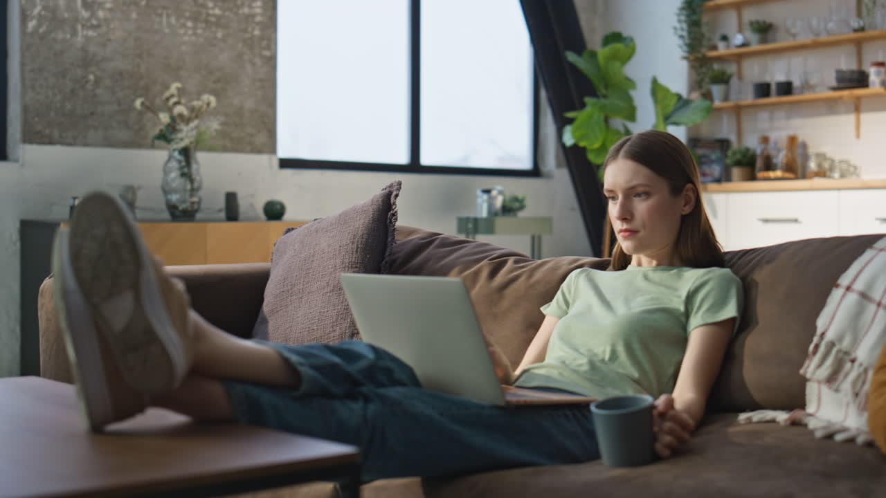 Domestic woman surfing laptop internet at home couch. Calm girl enjoying coffee