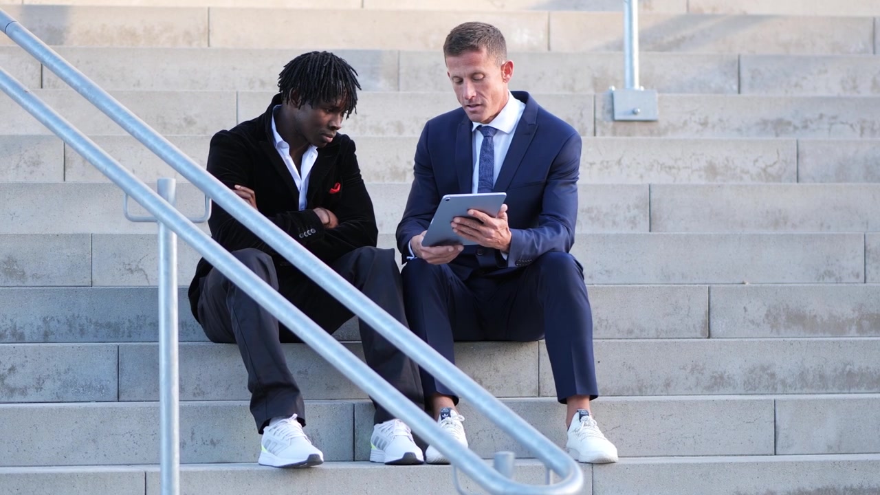 Orbit shot of two businessmen engaged in discussion while seated on stairs with tablet in hand