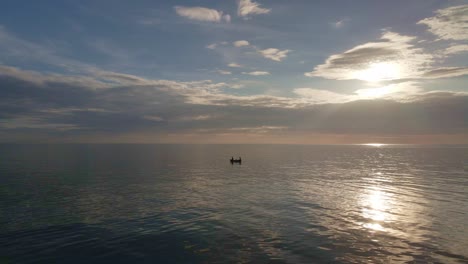 Aerial-drone-orbit-shot-of-ocean-silhouetted-at-sunset-with-small-fishing-boat-and-reflection-on-the-sea