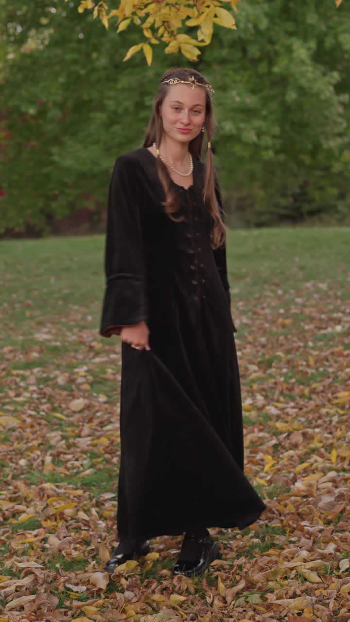 Vertical cinematic shot of a smiling medieval woman in black velvet dress turning gracefully and showing her crown under golden autumn leaves in warm natural light