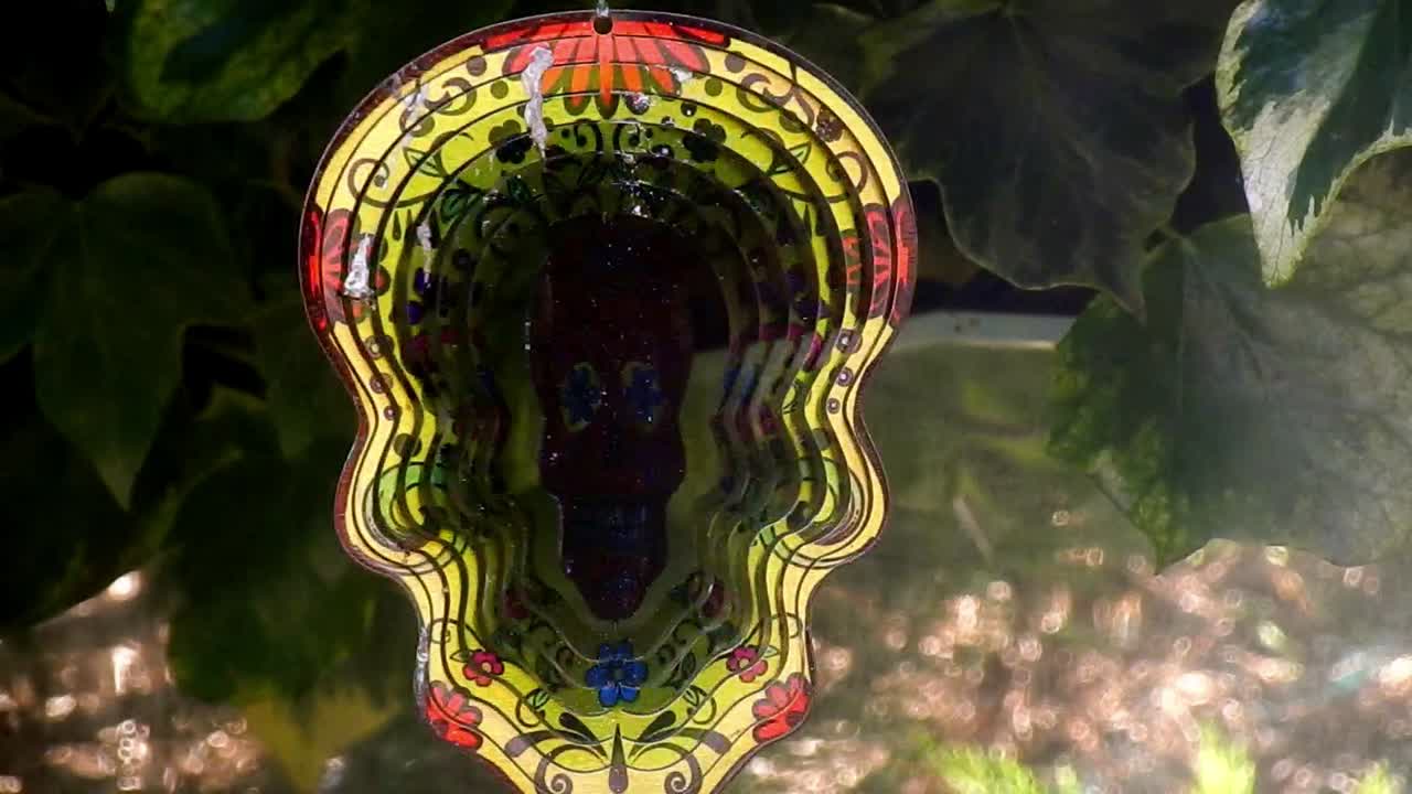 Ipnotico ornamento da giardino girevole, un teschio di zucchero in metallo riccamente decorato e colorato, appeso a un albero.