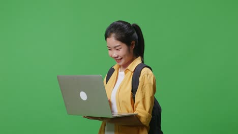 side view of asian woman student with a backpack smiling and typing on a laptop while standing in the green screen background studio