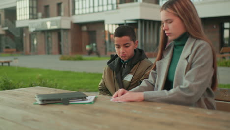 Male child attentively watches aunt folding paper into plane while sitting outdoors at wooden table with notebooks, both focused on creative activity in urban environment near residential buildings