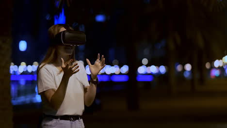 A-young-woman-in-virtual-reality-glasses-in-the-night-city-moves-her-hands-mastering-the-application-interface.-VR-technologies-of-the-future-in-everyday-life.-Virtual-reality-games