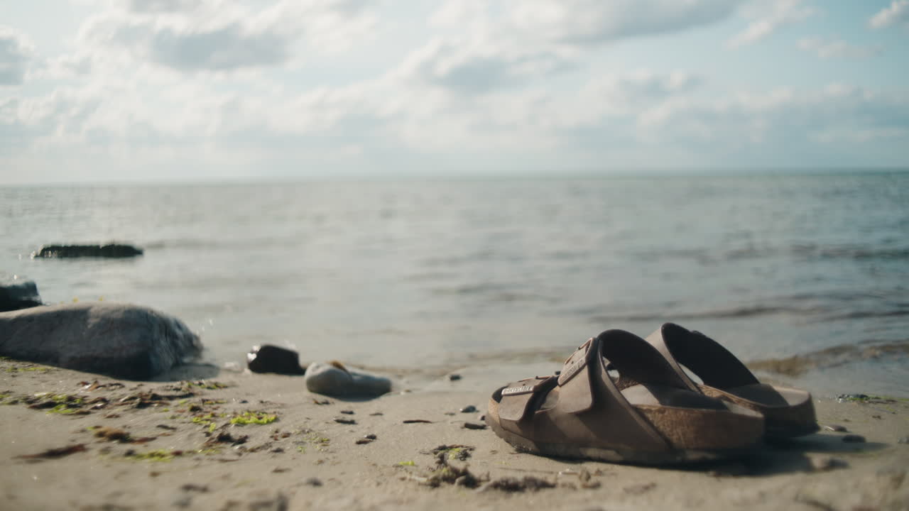 Women picks up sandals on stone beach along the ocean on a sunny day, Sandvik, &Ouml;land Sweden , close up wide