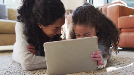 madre e hija de raza mixta felices acostadas en el suelo, divirtiéndose y usando la tableta