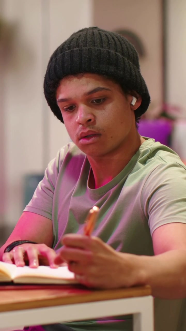 Vídeo vertical: Transição para homem focado à mesa após um olhar, escrevendo em caderno com caneta, gorro