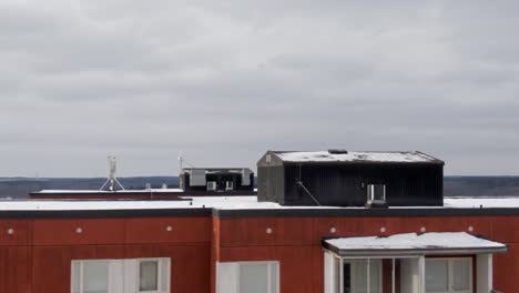 Drone-Footage-In-Akalla-Stockholm,-Sweden-Flying-Over-Red-Building-With-Black-Shed-On-Top-Of-Snow-Covered-Roof-Panning-To-Show-Winter-Snowy-Townscape-With-Smaller-Buildings-And-Traffic-On-The-Streets