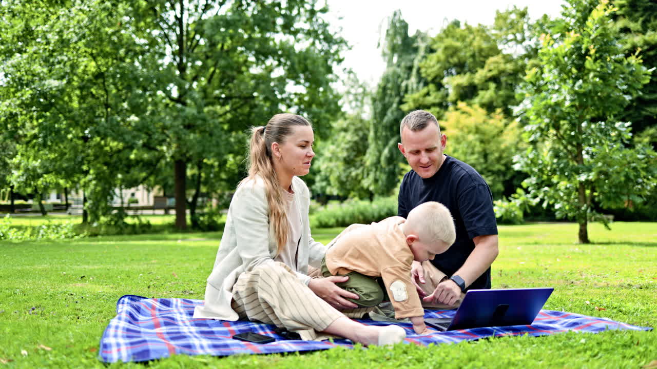 Familiepar med lille søn sidder i parken på græsset og ser video på laptop. Glad familie med barn og klapvogn udendørs. Mor, far og søn slapper af i naturen.