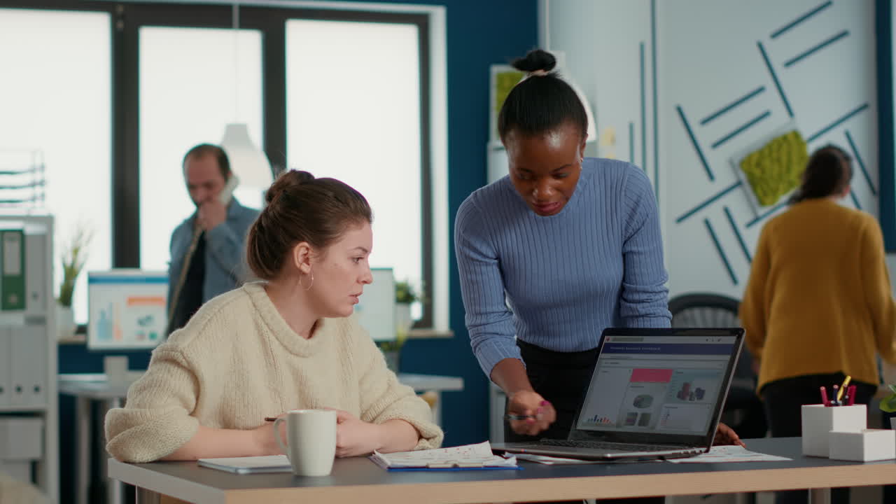 African american startup employee standing next to coworker presenting marketing strategy on laptop with charts