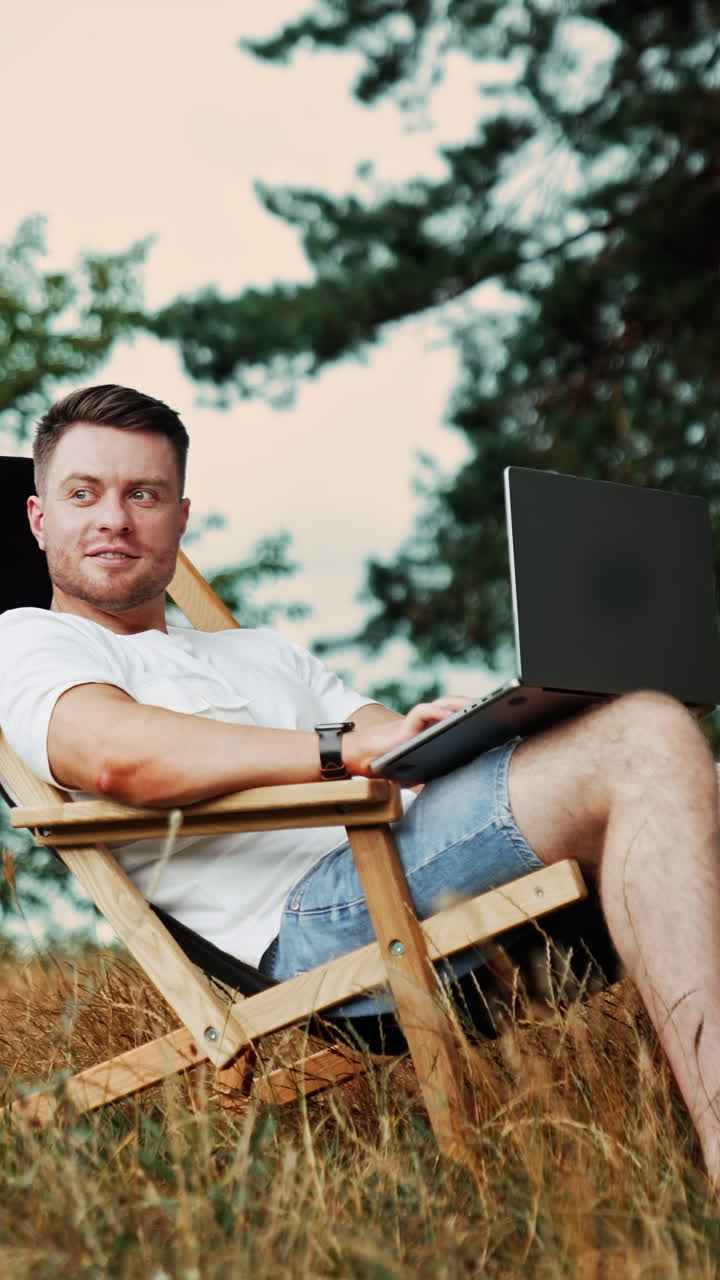 Positive smiling freelance male works in a comfortable chair. Remote work on the internet in the nature. Vertical video