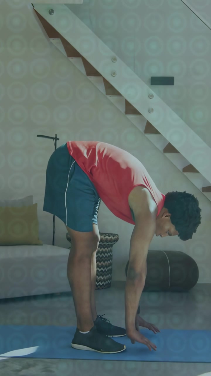 Vertical video: Man hinging hips to start forward bend on blue mat, lowering torso for fitness