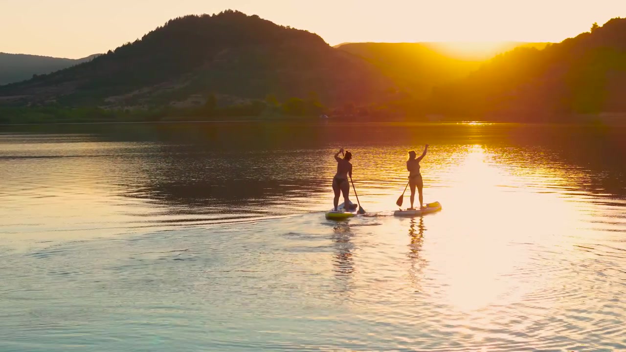 Due donne remano su un lago calmo al tramonto dorato con riflessi di montagna sull'acqua immobile, un tranquillo viaggio tra amiche, ripresa aerea orbitale