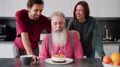 Un-Anciano-Feliz-Con-Cabello-Gris-Y-Una-Barba-Exuberante-Con-Una-Camisa-Rosa-Apaga-Una-Vela-En-Un-Pastel-Y-Sus-Hijos-Adultos,-Un-Hombre-Y-Una-Niña-Morena-Con-Una-Chaqueta-Verde,-Felicitan-A-Su-Papá-Por-Su-Cumpleaños-En-Un-Apartamento-Moderno.