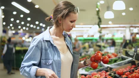 The-girl,-housewife-shopping-at-the-supermarket.-Chooses-fresh-tomatoes-on-a-branch,-puts-them-in-a-cellophane-bag,-ties-them-and-puts-them-into-a-cart.-Full-of-people-store.-Handheld