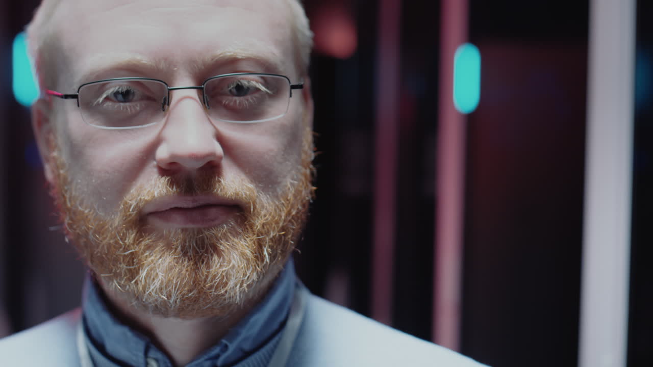 Close-up portrait of a man with red hair, beard, and glasses in a tech environment