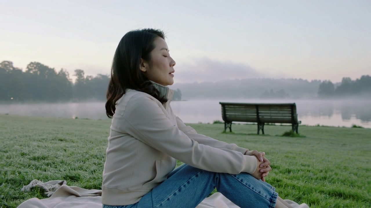 Ao amanhecer, mulher à esquerda ajustando as mãos, sentada à beira do lago na névoa sobre um cobertor, de jaqueta, meditando.