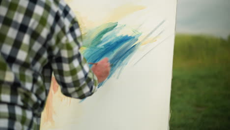 a back view of an illustrator wearing a black hat and checkered shirt, focused on painting a large, colorful canvas in a serene grass field