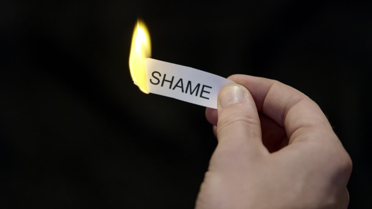 Bruciare via la vergogna - Una mano che dà fuoco a un biglietto di carta con la scritta “VERGOGNA”, una potente liberazione da critiche, sensi di colpa e vergogna. Primo piano cinematografico scuro. Salute mentale, terapia.